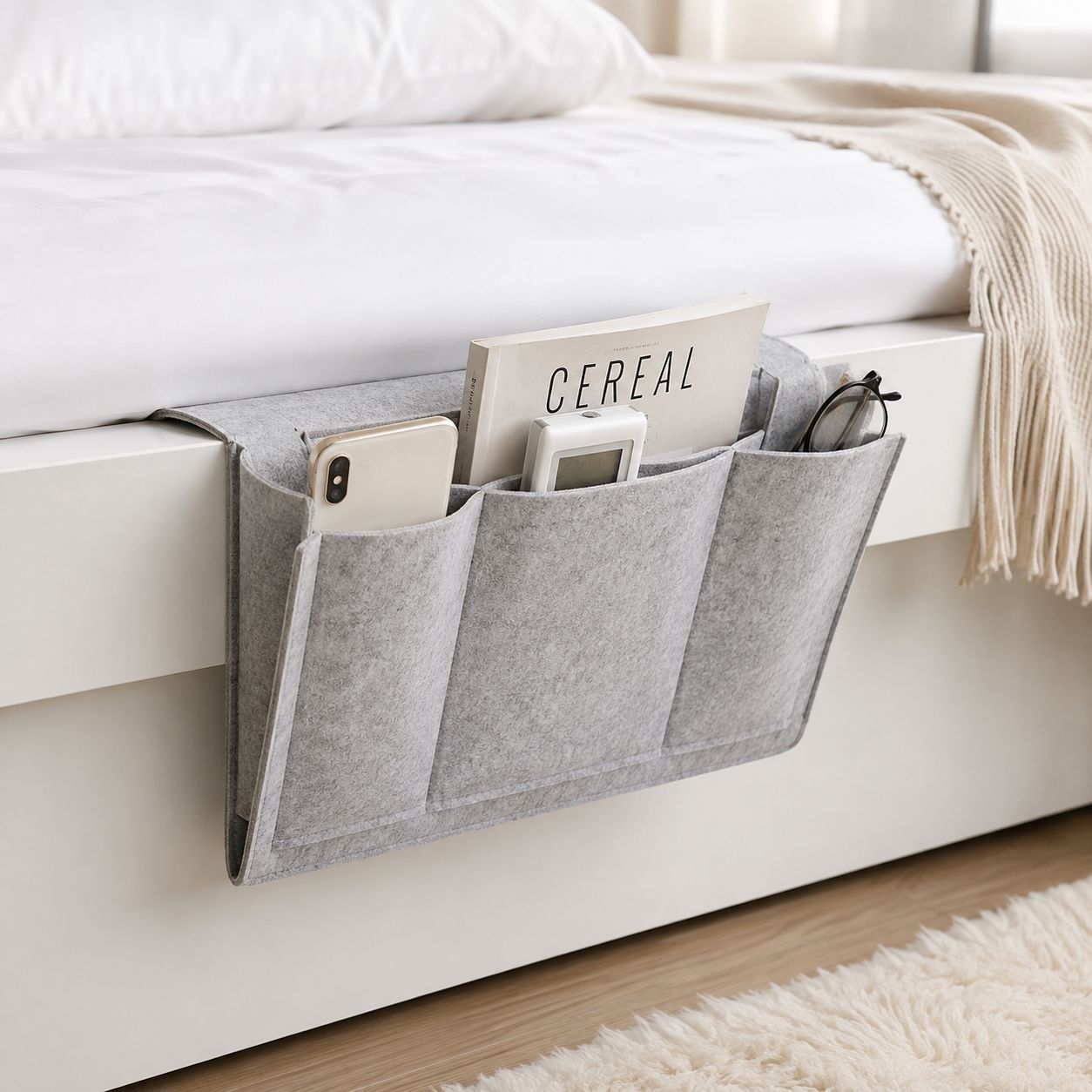 A light gray felt under-bed storage organizer with multiple pockets, shown hanging from the side of a white bed frame. The organizer is styled with everyday items like a smartphone, a cereal box, a small device, and eyeglasses, set against a clean bedroom background with white bedding and a beige throw blanket.