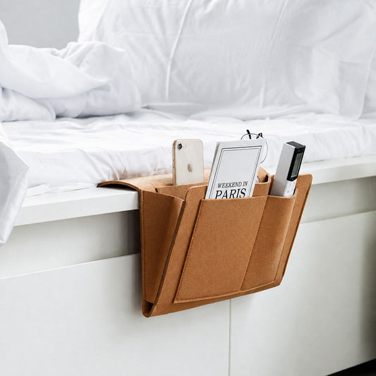 A brown felt under-bed storage organizer clipped to the edge of a white bed, holding a smartphone, a book, and a small black device.