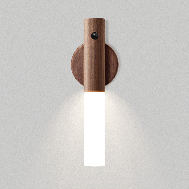 A modern wall-mounted light fixture with a cylindrical wooden upper section, a round wooden wall plate, and a glowing white vertical glass shade, shown against a light gray textured wall.