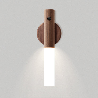 A modern wall-mounted light fixture with a cylindrical wooden upper section, a round wooden wall plate, and a glowing white vertical glass shade, shown against a light gray textured wall.